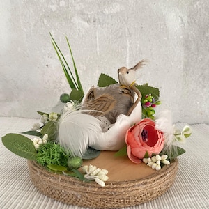 May include: A white egg with a bird sitting on top of it. The egg is surrounded by green leaves, white feathers, and a pink flower. The egg is sitting on a wooden base with burlap around the edge.