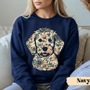 May include: Navy blue sweatshirt featuring a floral dog design. The dog's face is composed of beige, blue, and peach-colored flowers and leaves. The word "Navy" is printed in a white font on a beige rectangle.