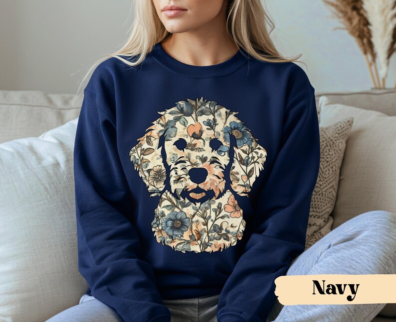 May include: Navy blue sweatshirt featuring a floral dog design. The dog's face is composed of beige, blue, and peach-colored flowers and leaves. The word "Navy" is printed in a white font on a beige rectangle.
