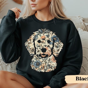 May include: Black sweatshirt featuring a floral dog design. The dog's face is composed of various flowers in shades of blue, peach, and cream, set against a cream background. The sweatshirt is a solid black color. The word "Black" is printed on a tan rectangle.