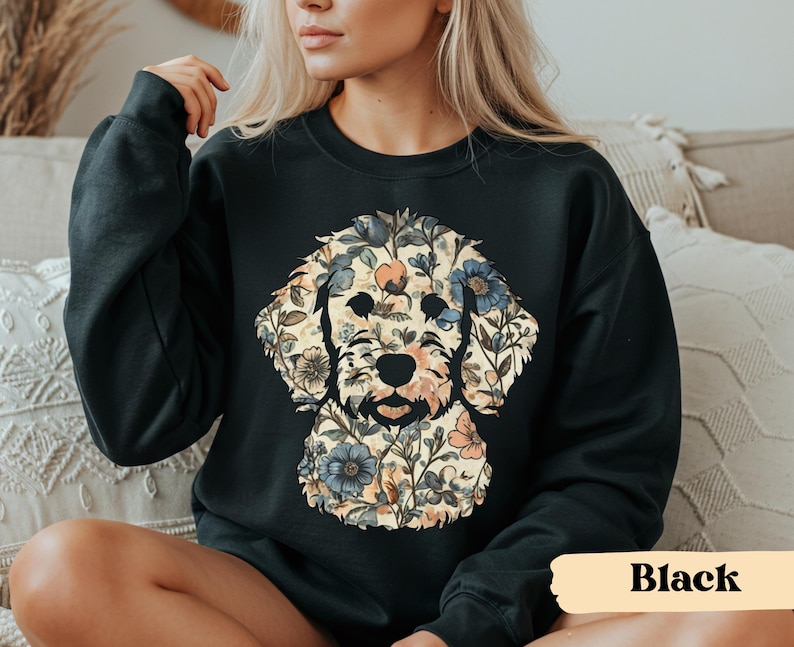 May include: Black sweatshirt featuring a floral dog design. The dog's face is composed of various flowers in shades of blue, peach, and cream, set against a cream background. The sweatshirt is a solid black color. The word "Black" is printed on a tan rectangle.