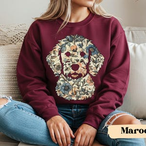 May include: Maroon sweatshirt featuring a floral dog design. The dog's face is composed of various flowers in shades of blue, peach, and cream. The sweatshirt is a solid maroon color, with the word "Maroon" in a tan banner at the bottom.