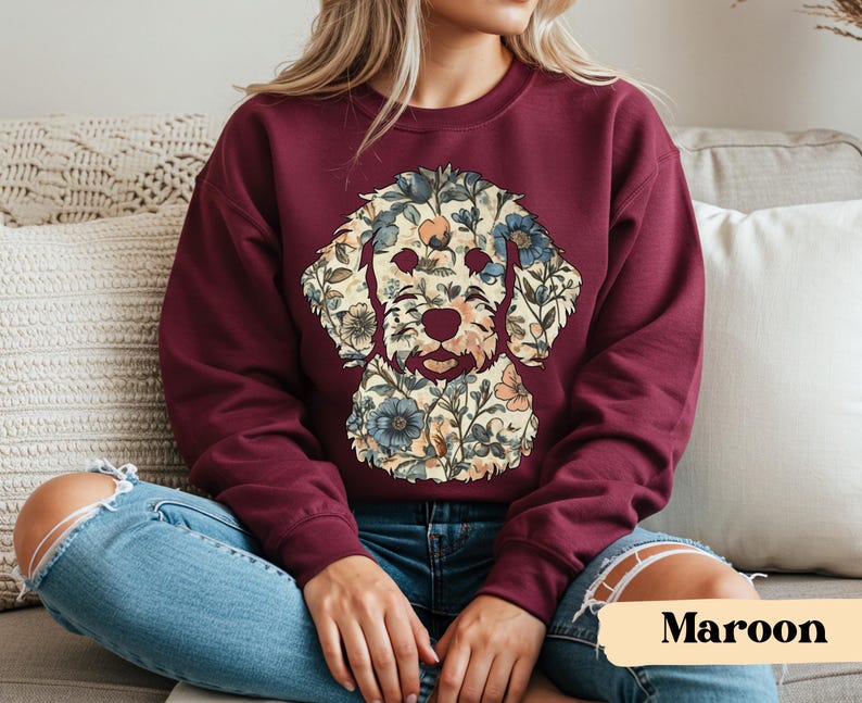 May include: Maroon sweatshirt featuring a floral dog design. The dog's face is composed of various flowers in shades of blue, peach, and cream. The sweatshirt is a solid maroon color, with the word "Maroon" in a tan banner at the bottom.