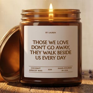 Puede incluir: Una vela encendida en un frasco de vidrio ámbar con tapa de metal. La etiqueta blanca presenta el texto: BY LAUREN, THOSE WE LOVE DON'T GO AWAY, THEY WALK BESIDE US EVERY DAY, COCONUT APRICOT WAX, 50H, HAND-POURED IN USA. La vela emite un brillo.