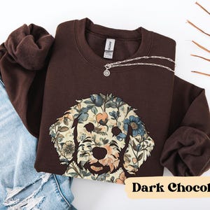 May include: A dark chocolate-colored sweatshirt featuring a floral dog face design. The dog's face is composed of blue, beige, and peach-colored flowers. A silver necklace with a circular pendant rests on the sweatshirt. The text "Dark Chocolate" is at the bottom.