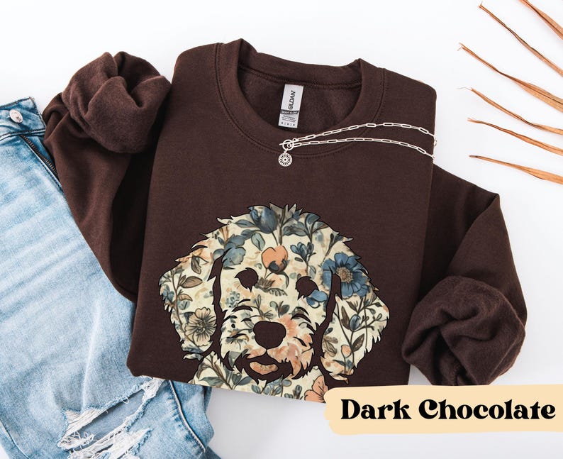 May include: A dark chocolate-colored sweatshirt featuring a floral dog face design. The dog's face is composed of blue, beige, and peach-colored flowers. A silver necklace with a circular pendant rests on the sweatshirt. The text "Dark Chocolate" is at the bottom.
