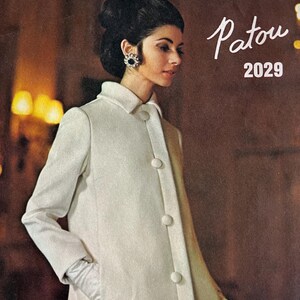 May include: A vintage Vogue Paris Original sewing pattern envelope featuring a white coat with a collar, long sleeves, and five large round buttons down the front. The text reads Vogue Paris Original, Patou, and 2029. A person is shown wearing the coat.