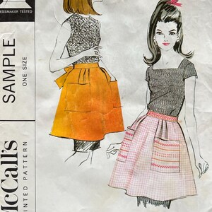 May include: A vintage McCall's sewing pattern envelope for a misses' and junior short apron. The front features illustrations of two apron styles, one solid orange with pockets and one pink gingham with decorative trim and pockets. Text includes SAMPLE.