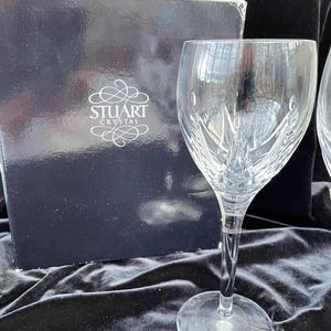 May include: Two clear glass wine glasses with a cut crystal design. The glasses are on a black surface with a black box that says "Stuart Crystal" on it.