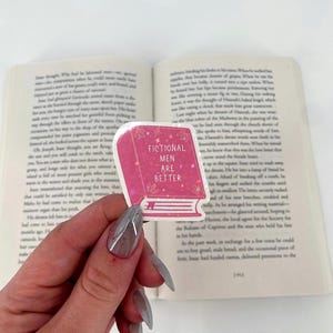 May include: A pink sticker in the shape of a book with the text "FICTIONAL MEN ARE BETTER" in white. The sticker is held in front of an open book with printed text.