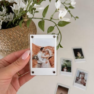 May include: A clear rectangular acrylic photo frame with four small magnetic corner fasteners holding a small rectangular photograph of a couple embracing in a desert landscape. The frame is held upright, displaying the photo clearly.