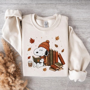 May include: A cream-colored crewneck sweatshirt featuring a graphic of Snoopy wearing a brown knit hat and scarf, reading a book next to a stack of books and a coffee cup. The design includes scattered brown autumn leaves on a light background.