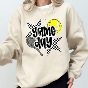 A woman wearing a beige sweatshirt with a graphic design featuring a tennis racket, a tennis ball, and the text "game day" printed on it.