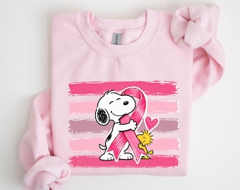 May include: A light pink crewneck sweatshirt featuring a graphic print of Snoopy hugging a large pink awareness ribbon with Woodstock standing nearby. A small pink heart is positioned above Woodstock, all set against horizontal brushstroke stripes.