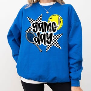 A woman wearing a blue sweatshirt with a graphic design featuring tennis equipment and the text "game day".