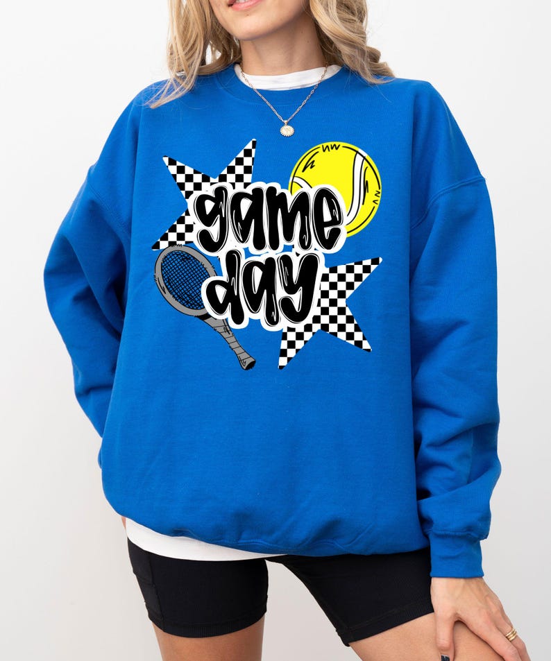 A woman wearing a blue sweatshirt with a graphic design featuring tennis equipment and the text "game day".