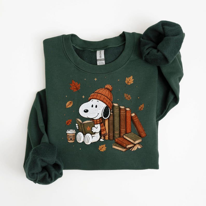 A green sweatshirt with a cartoon image of a dog wearing a hat and scarf, surrounded by books and autumn leaves.