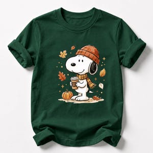 May include: A dark green short-sleeved t-shirt featuring a graphic of a white cartoon dog wearing an orange knit beanie and a plaid scarf. The dog holds a coffee cup, surrounded by falling autumn leaves and a small orange pumpkin on the ground.