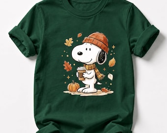 May include: A dark green short-sleeved t-shirt featuring a graphic of a white cartoon dog wearing an orange knit beanie and a plaid scarf. The dog holds a coffee cup, surrounded by falling autumn leaves and a small orange pumpkin on the ground.