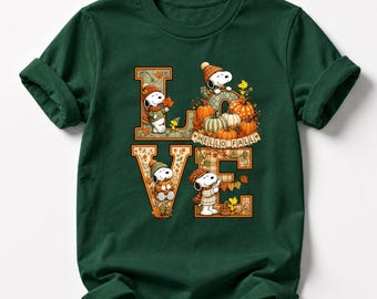 May include: A dark green short-sleeve t-shirt featuring a large graphic that spells the word LOVE. The letters are decorated with autumn-themed illustrations of Snoopy and Woodstock wearing hats and scarves, surrounded by pumpkins and fall leaves.