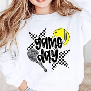 A smiling young woman wearing a white sweatshirt with a graphic design featuring a tennis racket and a tennis ball, along with the text "game day".