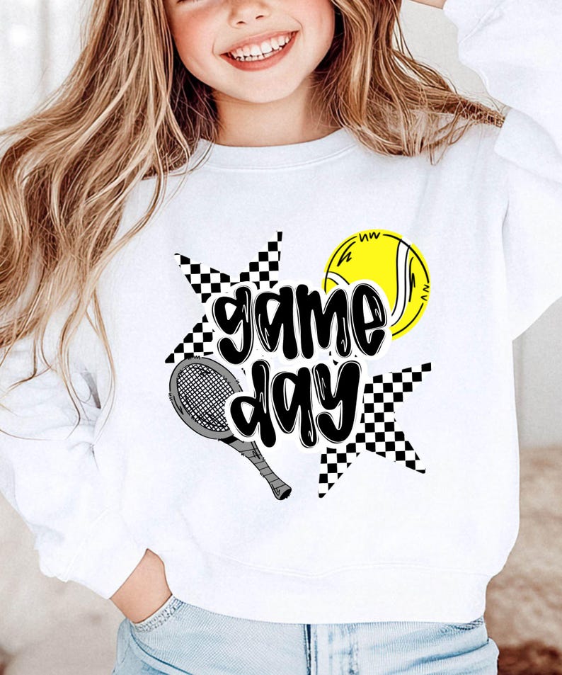 A smiling young woman wearing a white sweatshirt with a graphic design featuring a tennis racket and a tennis ball, along with the text "game day".
