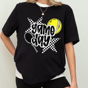 A woman wearing a black t-shirt with a graphic design featuring tennis equipment and the text "game day" printed on it.