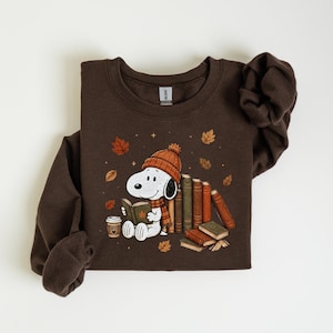 A brown sweatshirt with a cartoon image of a white dog wearing a hat and scarf, holding a book and surrounded by autumn leaves and books.