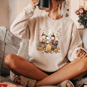 May include: A beige crewneck sweatshirt featuring a graphic print of the Peanuts characters sitting on a log around a campfire. The illustration includes Snoopy, Charlie Brown, Lucy, Linus, and Woodstock surrounded by autumn leaves and pumpkins.