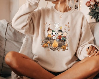 May include: A beige crewneck sweatshirt featuring a graphic print of the Peanuts characters sitting on a log around a campfire. The illustration includes Snoopy, Charlie Brown, Lucy, Linus, and Woodstock surrounded by autumn leaves and pumpkins.