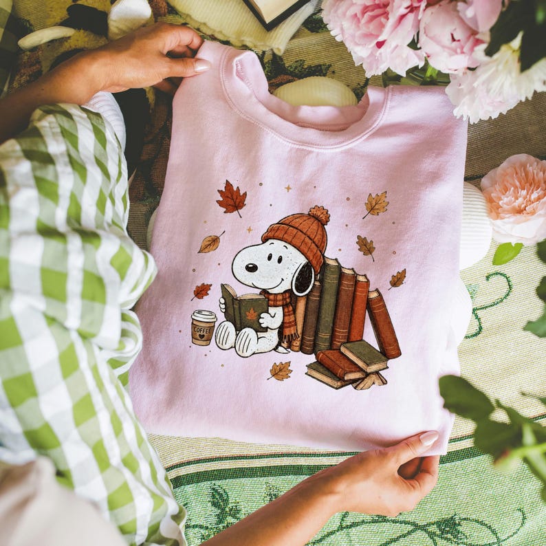 A person is holding a pink sweatshirt with a cartoon image of Snoopy, a character from the Peanuts comic strip, sitting with books and autumn leaves.