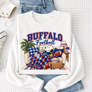 May include: A white long-sleeve sweatshirt featuring a graphic of Snoopy sitting on a couch under a blanket with the text BUFFALO Football. The design includes a blue and white plaid blanket, a bowl of popcorn, two footballs, and a lamp.
