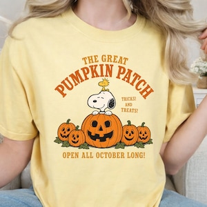 May include: A pale yellow t-shirt featuring a graphic of Snoopy sitting on a large jack-o'-lantern surrounded by smaller pumpkins. The text reads THE GREAT PUMPKIN PATCH, TRICKS! AND TREATS!, and OPEN ALL OCTOBER LONG! in orange lettering.