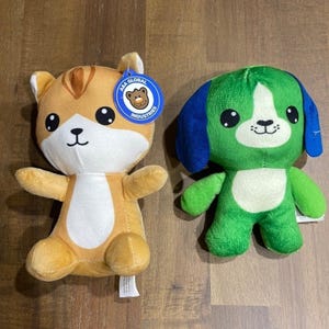 May include: Two plush toys: a tan and white cat with a blue circular tag reading A&A Global Industries and a green and blue dog. Both toys feature large black eyes, embroidered facial details, and a soft texture, standing upright on a flat surface.