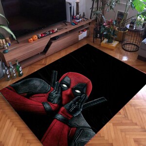May include: Black rectangular rug featuring a vibrant image of Deadpool in his red and black costume. The rug showcases Deadpool's face and hands in a gesture, with two katanas visible. The rug is placed on a wooden floor.
