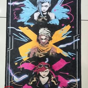 Pu&ograve; includere: Un tappetino per porta in tessuto nero con tre personaggi del videogioco League of Legends. I personaggi sono Jinx, Vi e Powder. I personaggi sono raffigurati in uno stile grafico colorato su uno sfondo nero.
