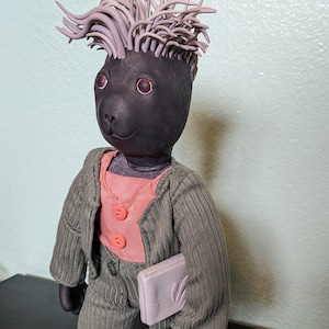 May include: A black plush toy with a pink shirt and a green corduroy suit. The toy has a purple spiky hairdo and is holding a pink clutch.