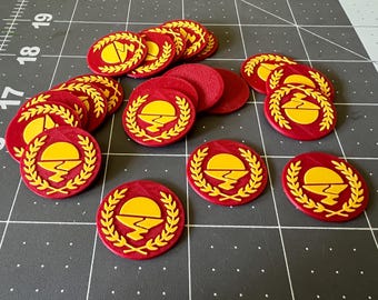 May include: A collection of circular red tokens featuring a raised yellow emblem of a sun rising over a jagged horizon, framed by a laurel wreath. The tokens are scattered across a dark gray cutting mat with visible white grid lines and numbers.