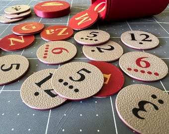 May include: A collection of circular tokens in red and beige, featuring printed numbers, letters, and dot patterns. Some tokens are plain red with gold letters, while others are textured beige with black or red numbers and corresponding dot counts.
