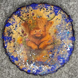 May include: A round wooden coaster with a rustic edge, featuring a Highland cow illustration. The cow is surrounded by flowers and glitter, with a blue and gold painted border. A decorative item.