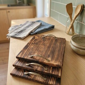 Puede incluir: Tres tablas de servir de madera con asas metálicas. Las tablas tienen un acabado oscuro y texturizado, apiladas sobre una superficie de madera clara. Ropa de cocina y utensilios en el fondo.