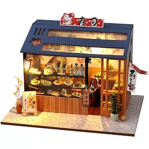 May include: Miniature sushi restaurant model with a blue roof, glass display, and wooden accents. The model features a sign with Japanese characters and a lucky cat figurine. The interior is lit, showcasing sushi and other Japanese food items.