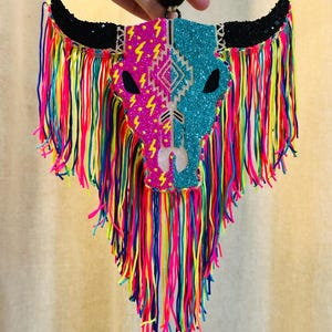 May include: A colorful bull skull wall hanging. The skull is split into pink and blue sections with glitter accents and a geometric design. Black horns and a fringe of rainbow-colored yarn complete the design.