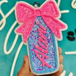 May include: A pink glitter bow-shaped ornament with a tag design. The tag is blue with pink text that reads "Mia". The ornament has a white border and a black metal loop for hanging.