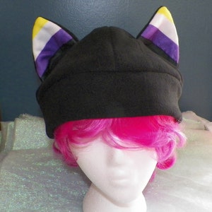 Puede incluir: Gorro de lana negro con orejas de gato amarillas, blancas, moradas y negras. El gorro está en una cabeza de maniquí con pelo rosa.