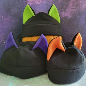 May include: Three black fleece cat ear hats with different colored ears. The largest hat has neon green ears, the middle hat has purple ears, and the smallest hat has orange ears.