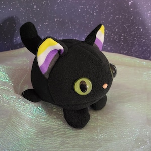May include: A black plush cat with large, round, green eyes and a small pink nose. The cat's ears are lined with the non-binary pride flag colors: yellow, white, purple, and black. The cat is a handmade item.