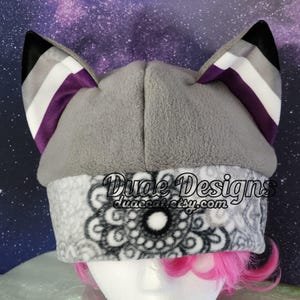 Puede incluir: Sombrero de orejas de gato de forro polar gris con un diseño de bandera de orgullo asexual de color negro, blanco y morado en las orejas. El sombrero tiene un forro polar estampado en blanco y negro. El texto "Duae Designs duaecat.etsy.com" está impreso en el sombrero.