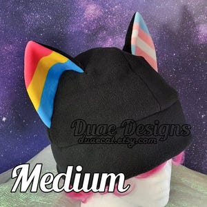 May include: Black beanie-style hat with colorful ear accents. One ear features the pansexual pride flag, the other the transgender pride flag. The hat has the text "Medium" and "Duae Designs" with a website address.