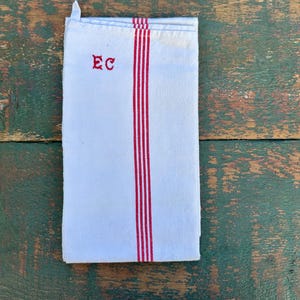 May include: White linen towel with three vertical red stripes and the embroidered letters "EC" in red. The towel is folded and has a small loop at the top. The towel is on a rustic wooden surface.